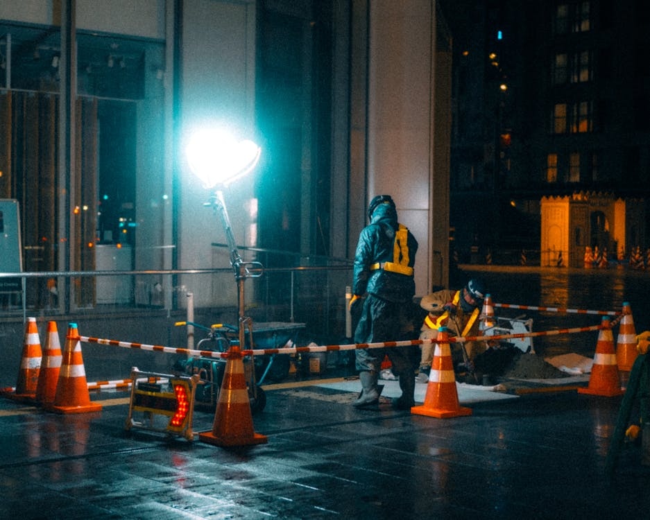 High-visibility orange traffic cones and safety barriers surrounding a road maintenance site under bright utility lighting