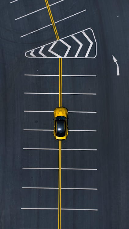 Aerial view of a yellow B2B service vehicle driving on a clean dark asphalt parking lot with double-yellow center lines and white parking slot grids